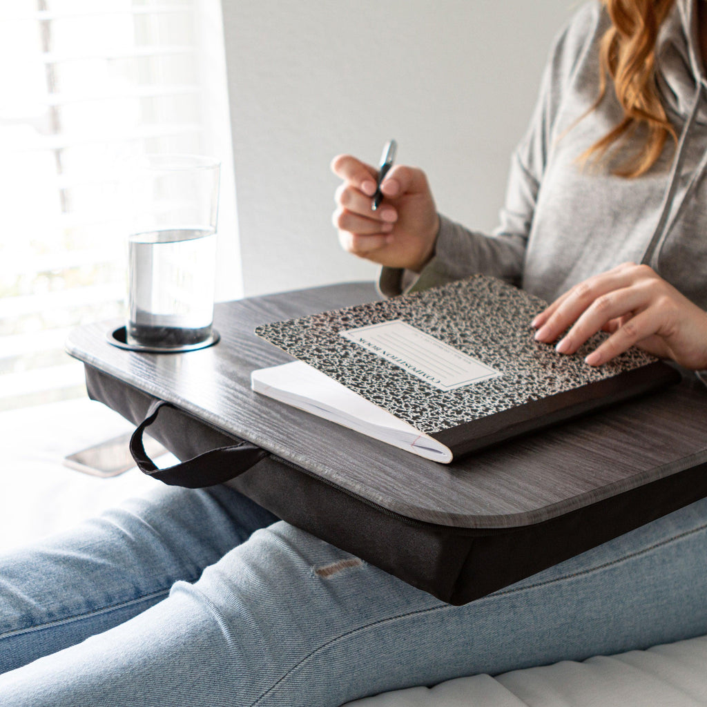 A woman sits on a bed with a lapdesk, writing in a notebook with a pen, surrounded by a cozy atmosphere.