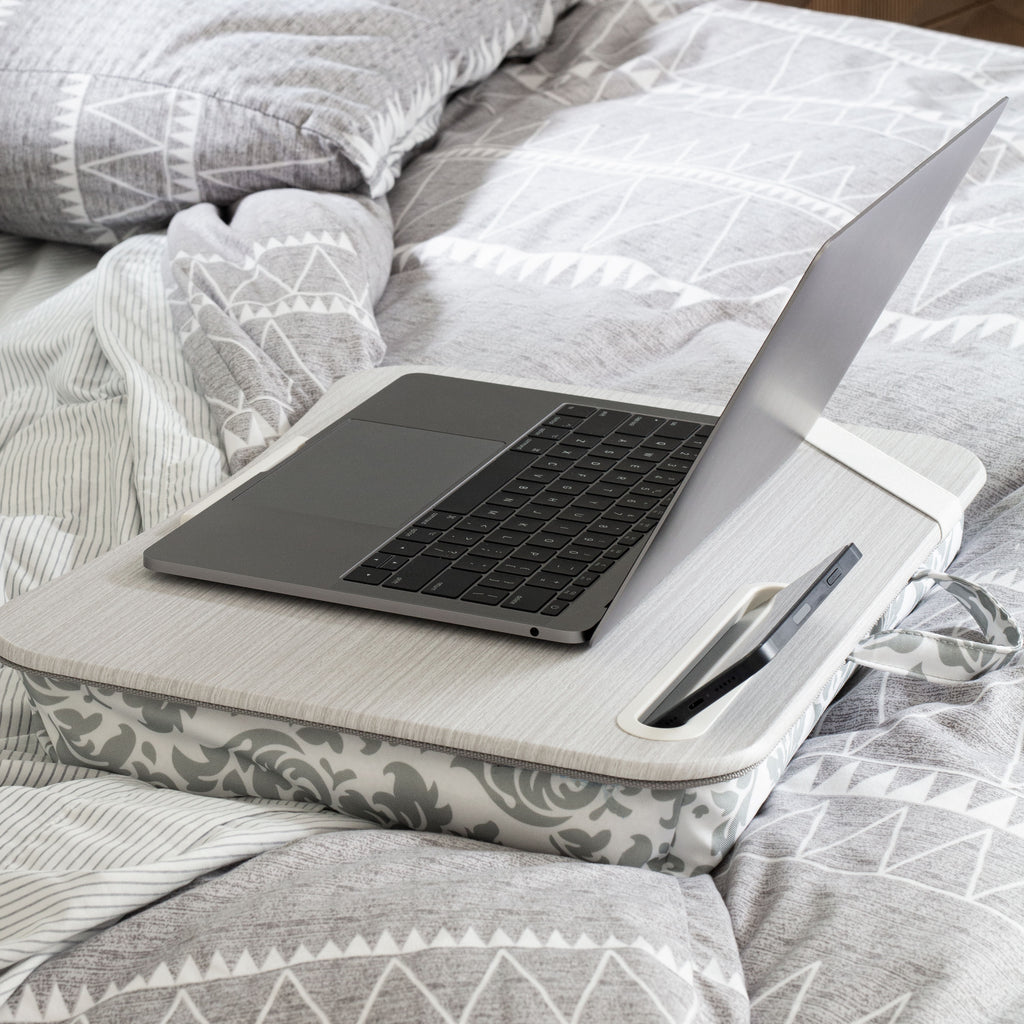 Gray Damask Designer Lap Desk is shown sitting on a bed with a laptop on its surface and a phone in the phone slot. 